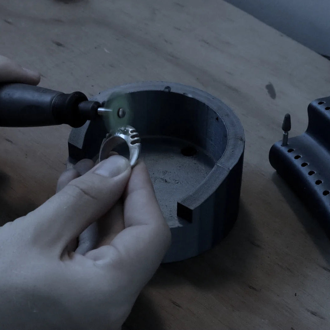A close-up of a person's hands using a rotary bursing tool to sharpen a small metal ring on a wooden workbench, with a ring stand holding multiple burr bits visible on the right.