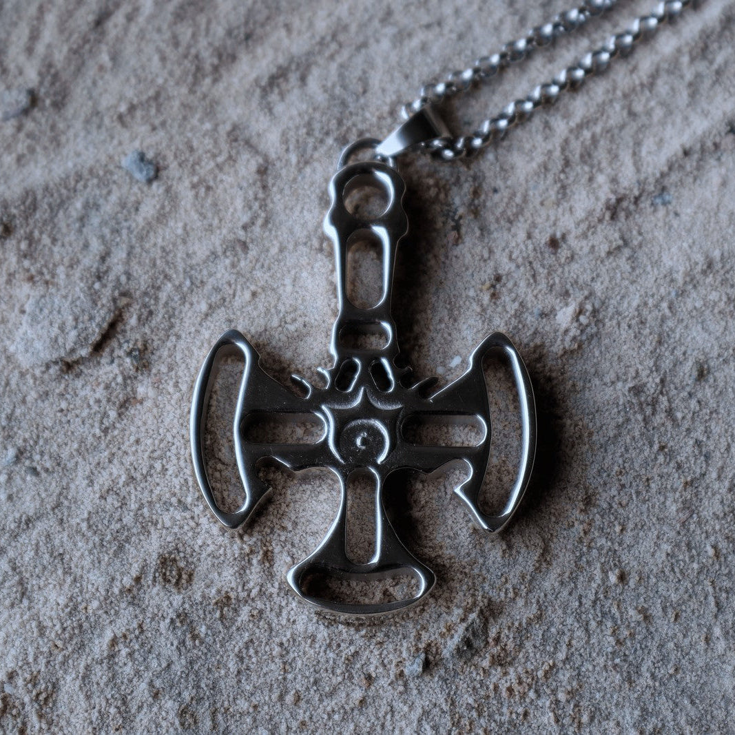 A silver-toned metal pendant necklace with a stylized cross design, resting on a textured sandy surface.