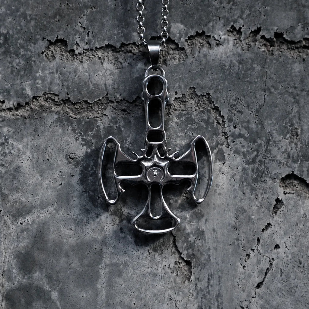A silver-toned metal pendant necklace featuring a stylized cross with axe-like details, hanging from a chain against a textured dark gray concrete background.