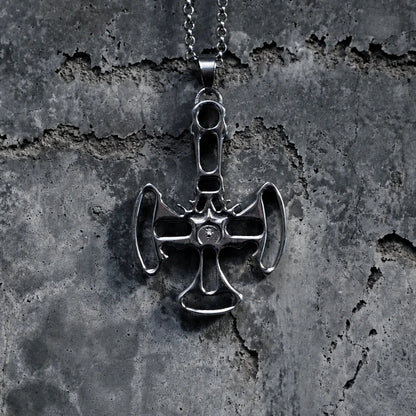 A silver-toned metal pendant necklace featuring a stylized cross with axe-like details, hanging from a chain against a textured dark gray concrete background.