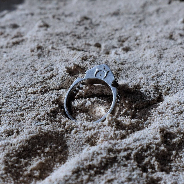 A silver-toned ring with a starburst and crescent moon design sits partially buried in light beige sand.