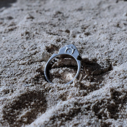 A silver-toned ring with a starburst and crescent moon design sits partially buried in light beige sand.