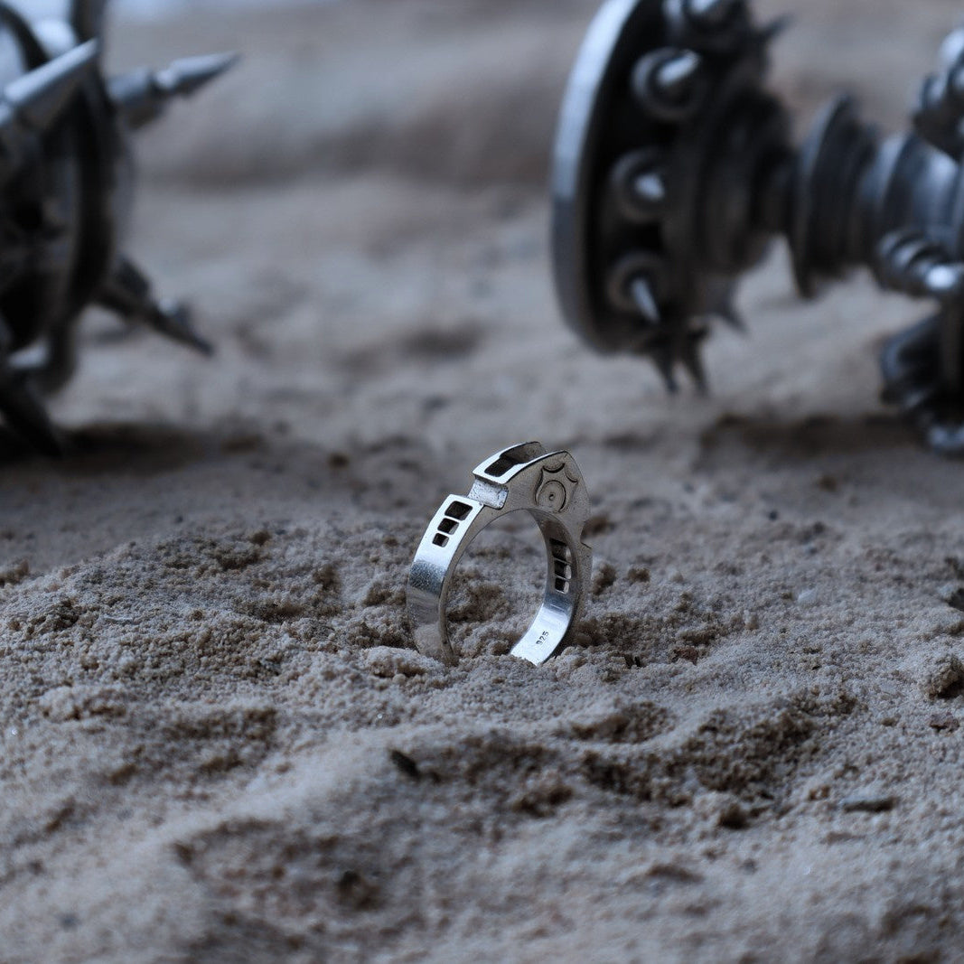 A silver ring with engraved details stands upright in sandy ground, with blurred mechanical spiky objects in the background.