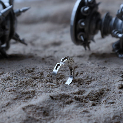 A silver ring with engraved details stands upright in sandy ground, with blurred mechanical spiky objects in the background.