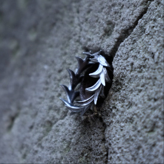 A silver-toned leaf-shaped ring with a textured, hammered finish, shown nestled in a crevice of a rough gray stone wall.